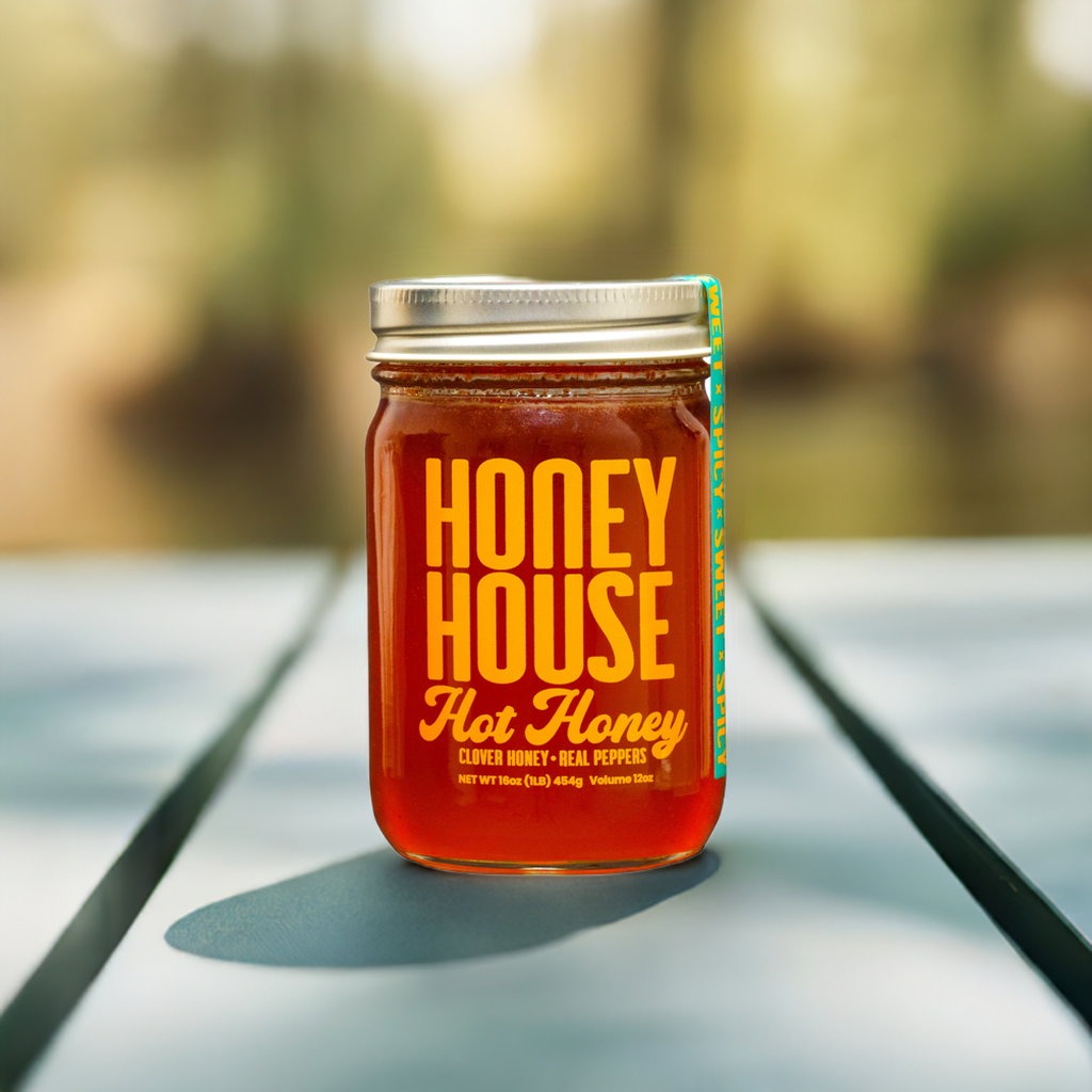 A jar of Honey House Honey Hot Honey filled with golden, raw honey sits on a sunlit wooden surface, promising a touch of natural heat against a softly blurred outdoor backdrop.