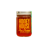 A glass jar of Honey House Honey’s Hot Honey, featuring red-orange honey and a silver lid, sits against a black background. The label notes clover honey, real peppers for heat, and a net weight of 454g.