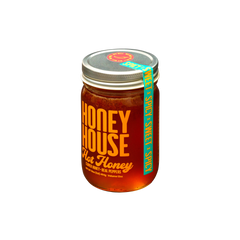 A glass jar of Honey House Honey Hot Honey, featuring orange and yellow text, contains raw honey infused with real peppers for natural heat. The jar has a metal lid and is set against a black background.